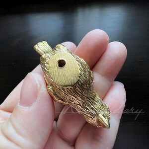 Puede incluir: Una figurita de p&aacute;jaro de metal de color dorado con una superficie texturizada detallada. El p&aacute;jaro est&aacute; mirando hacia abajo con las alas extendidas. Hay un peque&ntilde;o agujero en la parte inferior de la figurita.
