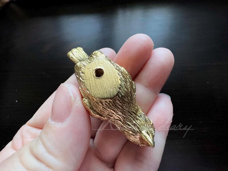 Puede incluir: Una figurita de p&aacute;jaro de metal de color dorado con una superficie texturizada detallada. El p&aacute;jaro est&aacute; mirando hacia abajo con las alas extendidas. Hay un peque&ntilde;o agujero en la parte inferior de la figurita.