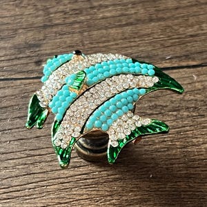 Può includere: Spilla decorativa a forma di pesce con base dorata. Il pesce presenta perline turchesi, dettagli in cristallo trasparente e pinne in smalto verde. La spilla è appoggiata su una superficie di legno.