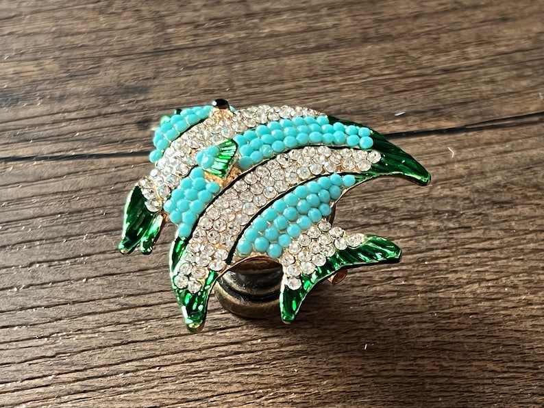 以下が含まれることがあります： 金色のベースに、ターコイズブルーのビーズ、クリアクリスタルのアクセント、グリーンのエナメルフィンの装飾的な魚の形をしたブローチ。ブローチは木製の表面に置かれています。