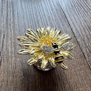 Könnte beinhalten: Eine dekorative Schmuckdose aus goldfarbenem Metall in Form einer Gänseblümchen mit einer Biene darauf. Die Biene hat schwarz-gelbe Streifen und kristallartige Akzente auf ihren Flügeln. Die Dose steht auf einer Holzoberfläche.