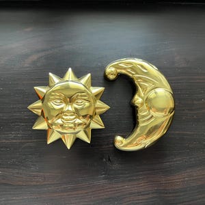 Pode incluir: Um conjunto de decoração de parede de sol e lua de cor dourada. O sol tem um rosto humano e a lua tem a forma de uma lua crescente.