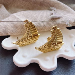 May include: Two gold-toned sailboat-shaped drawer pulls on a white ceramic base. The pulls have intricate details and a slightly raised design.