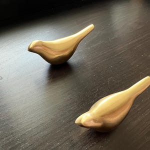Pu&ograve; includere: Due statuine di uccelli in metallo color oro. Gli uccelli sono stilizzati e astratti, con forme semplici e arrotondate. Sono entrambi rivolti verso il lato destro dell'immagine.