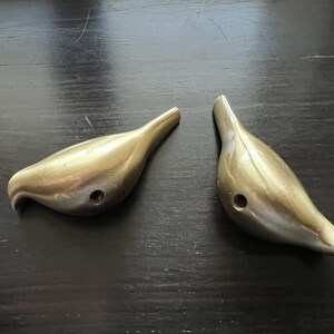 K&ouml;nnte beinhalten: Zwei goldfarbene Wandhaken in Vogelform. Die Haken sind aus Metall und haben eine glatte, gl&auml;nzende Oberfl&auml;che.