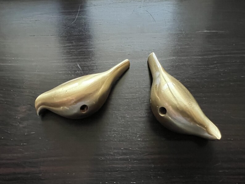 K&ouml;nnte beinhalten: Zwei goldfarbene Wandhaken in Vogelform. Die Haken sind aus Metall und haben eine glatte, gl&auml;nzende Oberfl&auml;che.