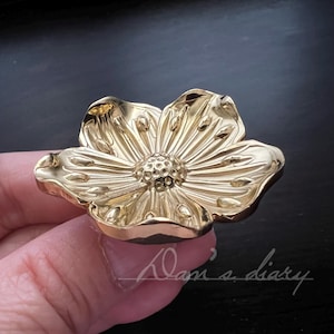 May include: A gold-coloured, flower-shaped cabinet knob. The knob has detailed petal and centre designs. The image is a close-up, showing the intricate details of the floral hardware. The background is dark.