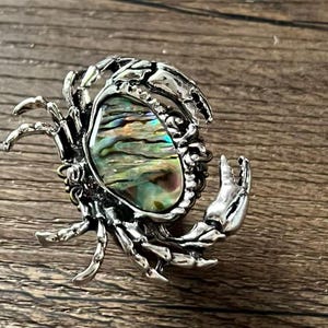 K&ouml;nnte beinhalten: Silberfarbene Krabbenbrosche mit einem gro&szlig;en, schillernden Abalone-Muschelzentrum. Die Krallen und Beine der Krabbe sind detailliert, und die Muschel zeigt eine Reihe von Farben, darunter Gr&uuml;n, Blau und Gold. Die Brosche liegt auf einer Holzoberfl&auml;che.