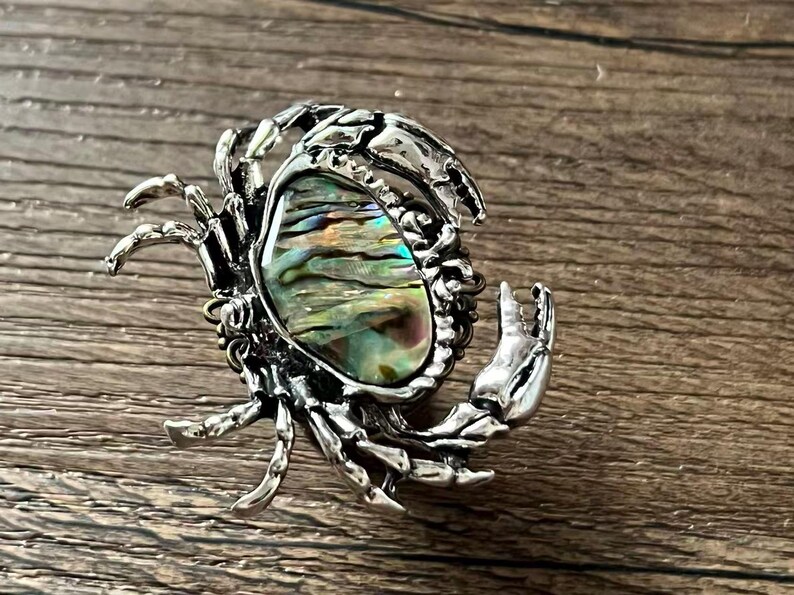 K&ouml;nnte beinhalten: Silberfarbene Krabbenbrosche mit einem gro&szlig;en, schillernden Abalone-Muschelzentrum. Die Krallen und Beine der Krabbe sind detailliert, und die Muschel zeigt eine Reihe von Farben, darunter Gr&uuml;n, Blau und Gold. Die Brosche liegt auf einer Holzoberfl&auml;che.