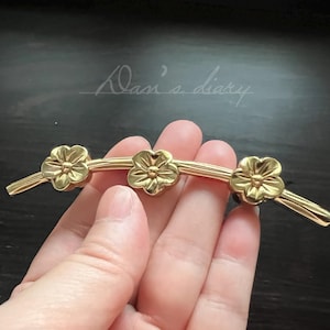 Peut inclure: Poignée de meuble dorée avec un motif floral. La quincaillerie comprend trois pièces en forme de fleur reliées par une barre métallique incurvée.