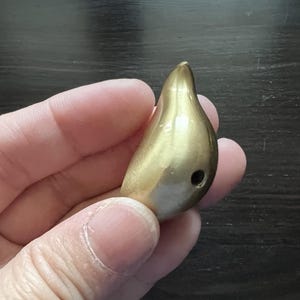 K&ouml;nnte beinhalten: Ein goldfarbener, tropfenf&ouml;rmiger Anh&auml;nger mit einem kleinen Loch am Boden.