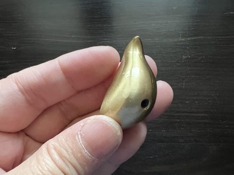 K&ouml;nnte beinhalten: Ein goldfarbener, tropfenf&ouml;rmiger Anh&auml;nger mit einem kleinen Loch am Boden.