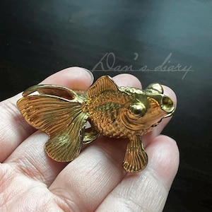 Könnte beinhalten: Eine goldfarbene Metallfigur eines Goldfisches. Die Figur ist mit Schuppen und Flossen detailliert.