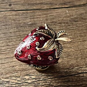 Op de afbeelding: Decoratieve aardbeivormige knop met een rode emaille afwerking en goudkleurige metalen accenten. De knop heeft een gedetailleerd ontwerp van bladeren en zaden. Ideaal voor meubels of decoratieve projecten.