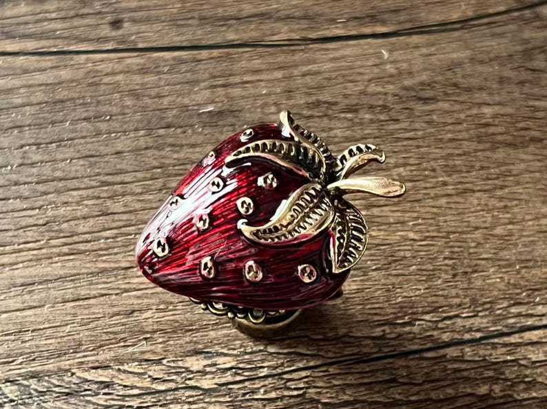 Op de afbeelding: Decoratieve aardbeivormige knop met een rode emaille afwerking en goudkleurige metalen accenten. De knop heeft een gedetailleerd ontwerp van bladeren en zaden. Ideaal voor meubels of decoratieve projecten.