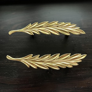 May include: Two gold-toned leaf-shaped drawer pulls. The pulls have a detailed design with a textured surface.