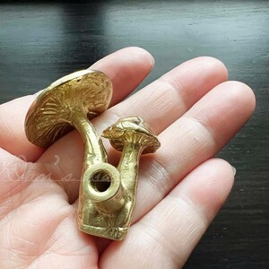 May include: Two gold-colored mushroom-shaped door knobs. The larger mushroom has a detailed cap and stem. The smaller mushroom is attached to the larger mushroom's stem.