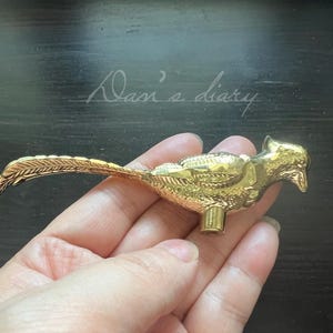 May include: A gold-colored bird-shaped decorative object. The bird has detailed feather and wing patterns, with a long tail and a small, cylindrical base. The object is held in a hand against a dark background. The text "Dan's diary" is visible in the background.