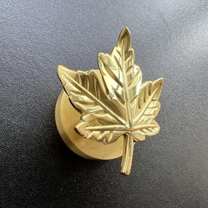Może przedstawiać: Złoty uchwyt do szafki w kształcie liścia klonu. Uchwyt ma szczegółowy wzór liścia i jest zamontowany na okrągłej podstawie. Tło stanowi teksturowana czarna powierzchnia.