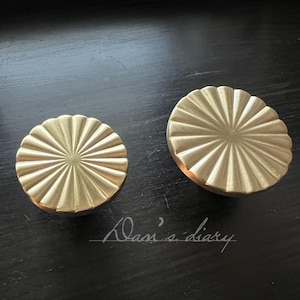 May include: Two gold-toned metal drawer pulls with a sunburst design. The pulls are round and have a raised, textured surface.