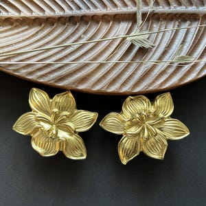 Pode incluir: Um par de brincos de flor dourados. Os brincos são feitos de metal e têm um design floral detalhado.