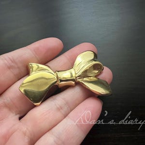 Può includere: Un fermaglio a forma di fiocco in metallo color oro. Il fiocco è annodato e ha una finitura liscia e lucida.