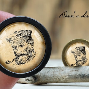 May include: Two round cabochons with a vintage illustration of a man with a beard and a cap smoking a pipe. One cabochon is black, the other is bronze. The text "Alan's diary" is written in cursive.