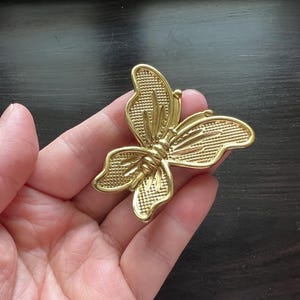 May include: A gold butterfly brooch with intricate wing detailing. The metal brooch has a textured finish. This classic accessory is suitable for adding a touch of elegance to any outfit. The brooch is approximately 6 cm wide.