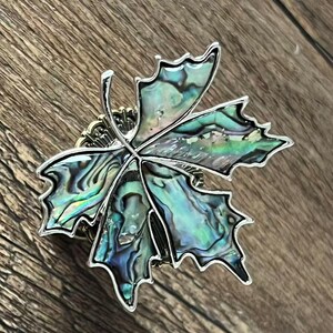 Peut inclure: Broche en forme de feuille avec un cadre en m&eacute;tal argent&eacute;. La feuille est remplie de nacre iris&eacute;e, affichant des nuances de vert, de bleu et de noir. La broche est pos&eacute;e sur un fond en bois.