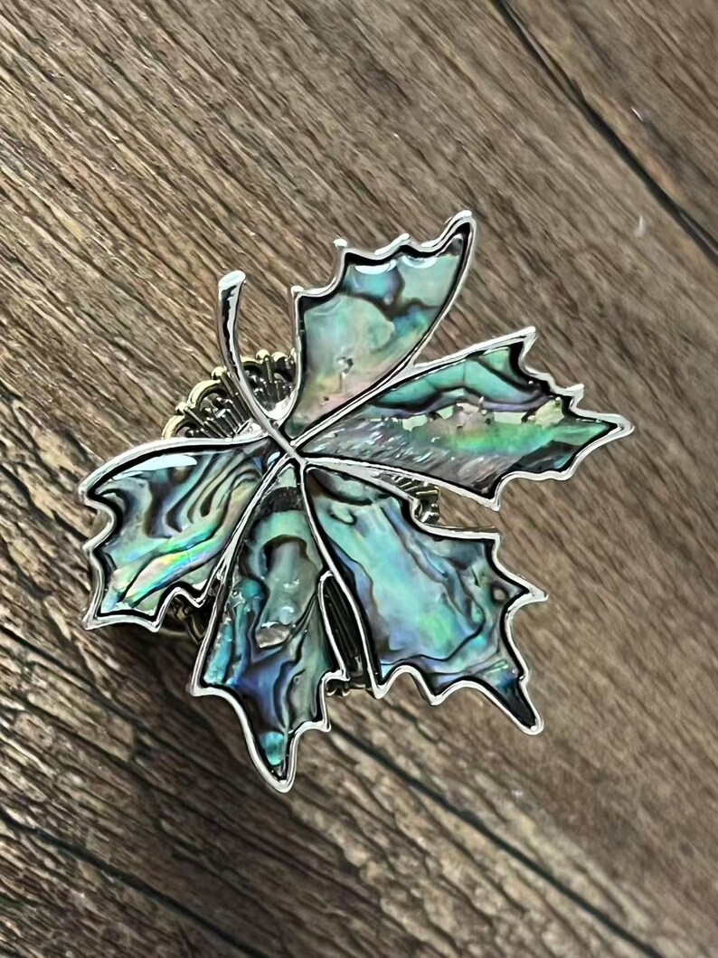 Peut inclure: Broche en forme de feuille avec un cadre en m&eacute;tal argent&eacute;. La feuille est remplie de nacre iris&eacute;e, affichant des nuances de vert, de bleu et de noir. La broche est pos&eacute;e sur un fond en bois.