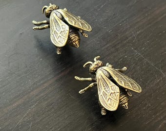 May include: Two brass-colored bee-shaped decorative objects. The bees have detailed wings, antennae, and legs. The objects are positioned on a dark, textured surface, suggesting a decorative or artistic theme.