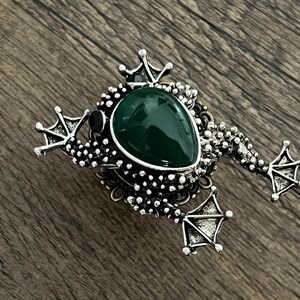 以下が含まれることがあります： 中央に大きな涙型の濃い緑色の石が付いた、銀色のカエルブローチ。 ブローチは、詳細な脚、王冠、テクスチャード加工されたボディが特徴です。 カエルは木製の表面に置かれています。