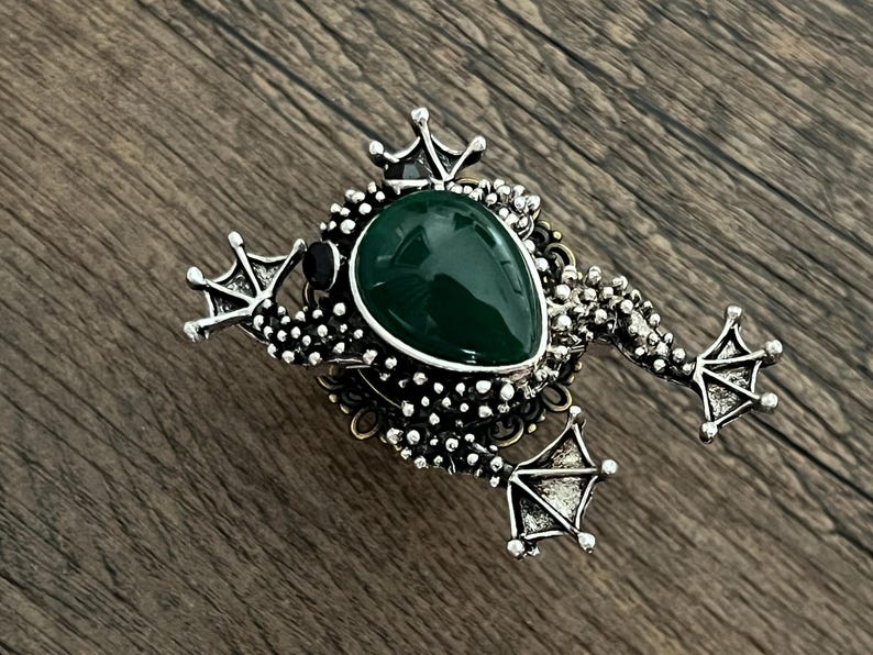 以下が含まれることがあります： 中央に大きな涙型の濃い緑色の石が付いた、銀色のカエルブローチ。 ブローチは、詳細な脚、王冠、テクスチャード加工されたボディが特徴です。 カエルは木製の表面に置かれています。