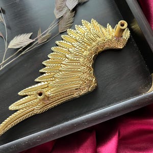 Op de afbeelding: Goudkleurige metalen vleugelvormige ladegreep met een gedetailleerd ontwerp. De greep heeft twee bevestigingsgaten aan de achterkant.