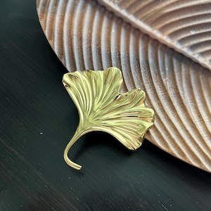 Op de afbeelding: Een goudkleurig decoratief object in de vorm van een ginkgoblad. Het blad heeft gedetailleerde nerven en een gebogen steel. Het ligt op een donkere ondergrond naast een houten, bladvormige dienblad.