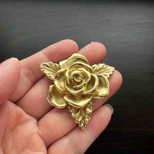 Peut inclure: Une broche en forme de rose dorée avec des pétales et des feuilles détaillés. Le motif floral est tenu dans une main sur un fond sombre. La broche est probablement en métal et a une esthétique vintage ou antique.