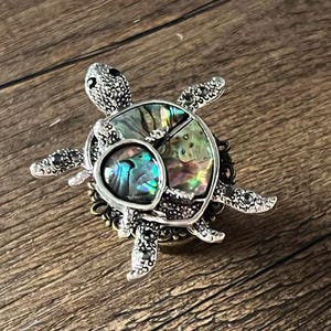 Op de afbeelding: Zilverkleurige schildpadbroche met een iriserende abalone schelp. De broche heeft gedetailleerde poten en een kop, geplaatst tegen een houten achtergrond. De schelp toont tinten groen, blauw en paars.