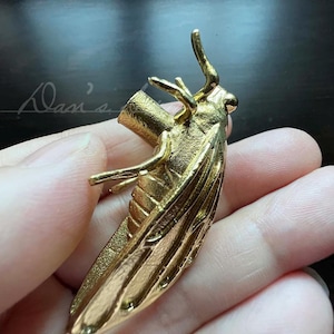 Può includere: Una statuetta di insetto in metallo dorato con ali dettagliate. La statuetta è tenuta in una mano.