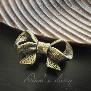 May include: A detailed, antique-style bow-shaped ornament in a metallic gold colour. The bow has a textured surface and is set against a dark background with a wooden, carved accent. The words "Dasi's diary" are written in cursive.