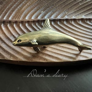 Könnte beinhalten: Eine Messing-Fischskulptur mit detaillierten Flossen und Schwanz liegt auf einem geschnitzten Holztablett in Blattform. Der Fisch hat ein schlankes, stromlinienförmiges Design. Das Tablett hat ein natürliches Holzmaserungsmuster. Der Text "Dani's diary" ist enthalten.