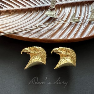 Pode incluir: Dois brincos de cabeça de águia de ouro. Os brincos são detalhados e têm um aspecto realista.