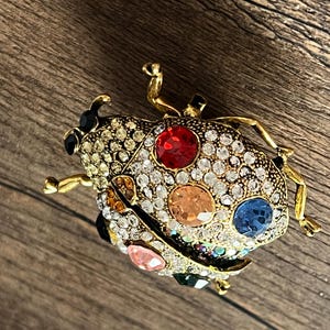May include: A gold-coloured ladybird brooch with a detailed design. The brooch features a body encrusted with clear rhinestones and colourful gemstones in red, orange, blue, pink, and green. The ladybird has black accents and gold-coloured legs and antennae.