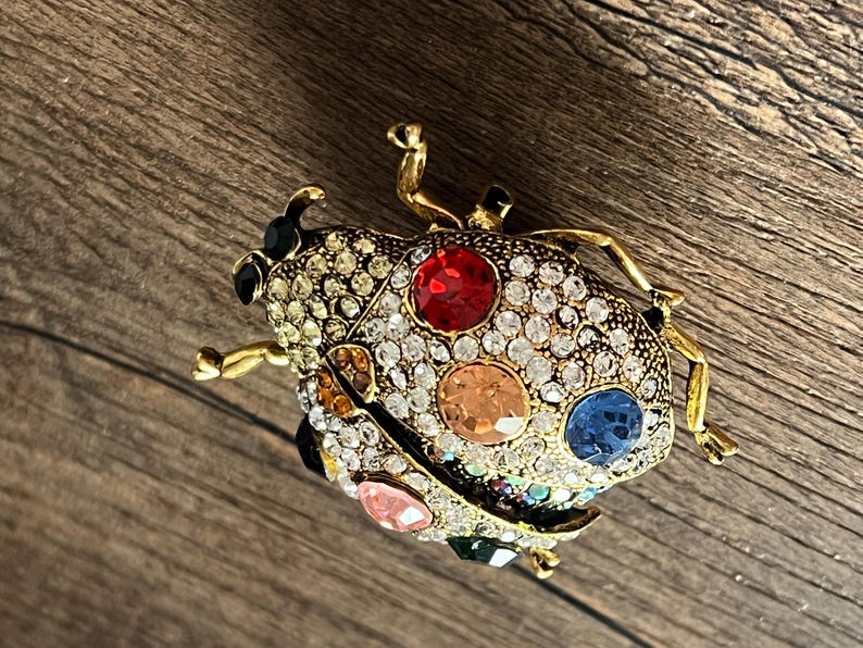 May include: A gold-coloured ladybird brooch with a detailed design. The brooch features a body encrusted with clear rhinestones and colourful gemstones in red, orange, blue, pink, and green. The ladybird has black accents and gold-coloured legs and antennae.