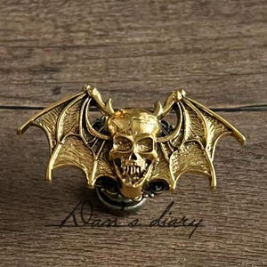 May include: A gold-colored skull pendant with bat wings and horns. The skull has detailed features and is mounted on a dark base. The wings are spread out, and the pendant is set against a wooden background.