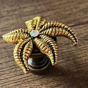 Op de afbeelding: Decoratieve kastknop in de vorm van een gouden zeester, met gedetailleerde ribbels en een iriserende edelsteen in het midden. De knop is gemonteerd op een donkere, metalen basis, geschikt voor meubels.