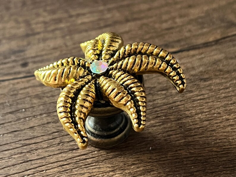 Op de afbeelding: Decoratieve kastknop in de vorm van een gouden zeester, met gedetailleerde ribbels en een iriserende edelsteen in het midden. De knop is gemonteerd op een donkere, metalen basis, geschikt voor meubels.