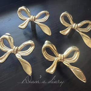 May include: Four gold-coloured bow-shaped cabinet pulls. The pulls are made of a shiny metal and are arranged on a dark surface. The pulls have a classic bow design, with a central knot and flowing loops.