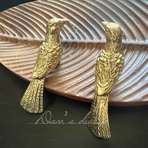 May include: Two gold-colored bird-shaped decorative objects. The birds are perched on a wooden leaf-shaped tray. The birds have detailed feather patterns and are positioned on a dark surface. The tray has a carved leaf design.