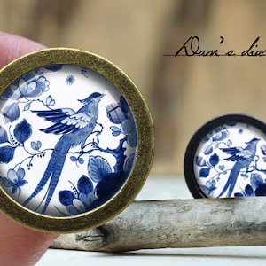 Op de afbeelding: Twee ronde kastknoppen met een blauw-wit vogel- en bloemmotief. De grotere knop heeft een bronzen rand, de kleinere een zwarte rand. De tekst "Dan's diary" is cursief geschreven.