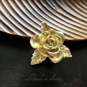 以下が含まれることがあります： 金色の金属製のバラで、花びらと葉が細かく作られています。バラは濃い茶色の木製の表面の上に置かれています。